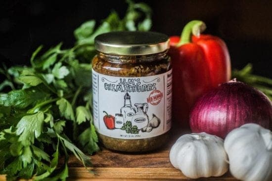 A jar of J&M's Chimichurri sauce with a label illustration of red pepper, oil, and vinegar, placed alongside fresh red bell pepper, a purple onion, garlic bulbs, and green parsley on a rustic wooden surface.