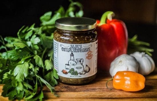 A jar of J&M's Chimichurri sauce, prominently displayed alongside lush green parsley, a vibrant red bell pepper, garlic bulbs, and an orange habanero pepper on a dark wooden backdrop.