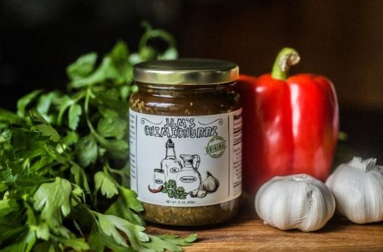 A jar of "J&M's Chimichurri" sauce surrounded by fresh parsley, a red bell pepper, and garlic bulbs on a wooden surface.