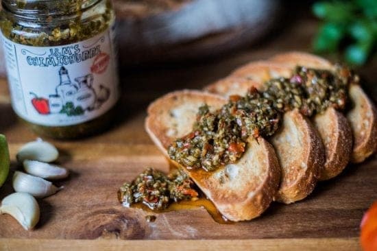 Toasted slices of bread generously topped with rich and flavorful chimichurri sauce, with a hint of the sauce jar in the background.